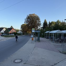 Fahrbahn links mit fahrradfahrender Person, mittig Bushaltestelle, rechts überdachte Fahrradparkplätze