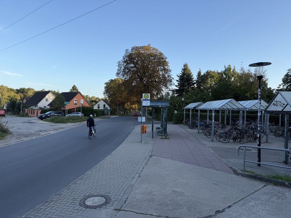 Fahrbahn links mit fahrradfahrender Person, mittig Bushaltestelle, rechts überdachte Fahrradparkplätze