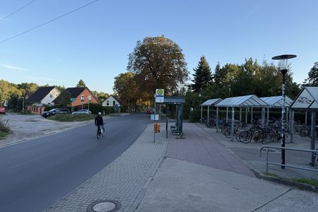 Fahrbahn links mit fahrradfahrender Person, mittig Bushaltestelle, rechts überdachte Fahrradparkplätze
