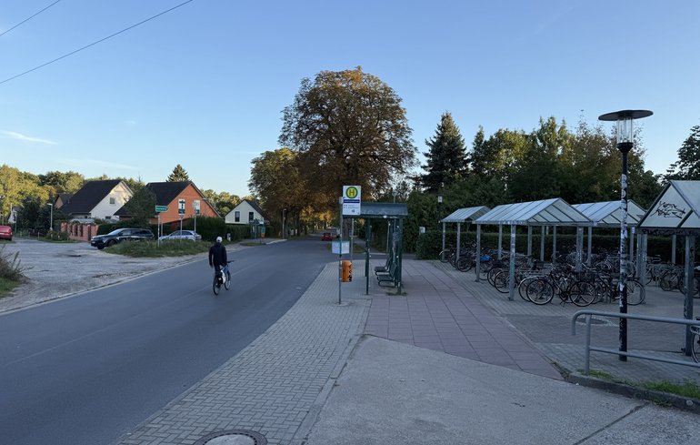 Fahrbahn links mit fahrradfahrender Person, mittig Bushaltestelle, rechts überdachte Fahrradparkplätze